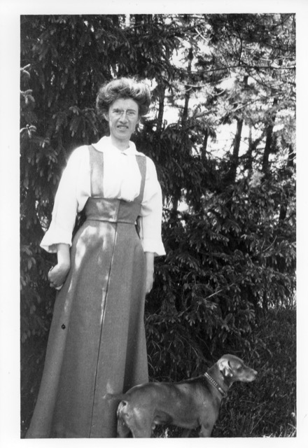 Sur la première image en noir et blanc, on voit Isabella Preston qui pose devant une rangée de conifères. Elle porte un robe-tablier avec une chemise à manches longues. Un chien prend la pose devant elle.