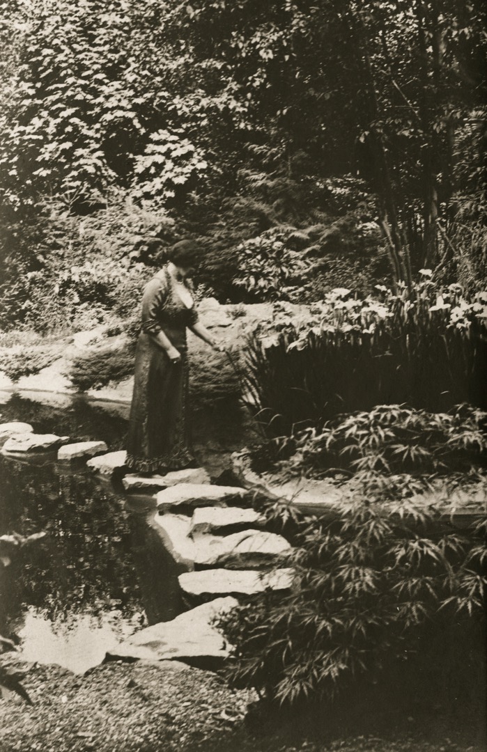 Sur cette photographie en noir et blanc, Jennie Butchart se promène dans son jardin. Elle se tient sur l’un des pavés du sentier qui traverse le ruisseau. À l’arrière-plan, on aperçoit des arbres feuillus. Au premier plan, des arbustes de différentes tailles bordent le sentier et le ruisseau.
