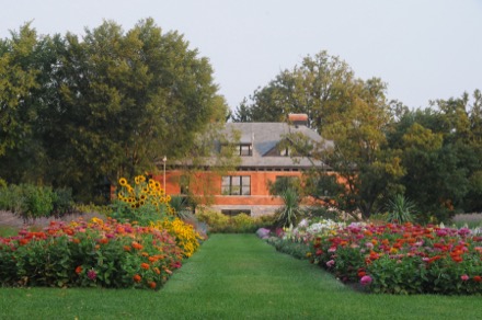 Sur la photographie couleur, on voit un bâtiment en briques rouges à l’arrière-plan. Cet édifice à deux étages occupe le centre de l’image. Un sentier gazonné mène jusqu’à lui. Deux platebandes fournies de plantes colorées bordent le sentier central. Des arbres feuillus entourent le bâtiment, au fond.