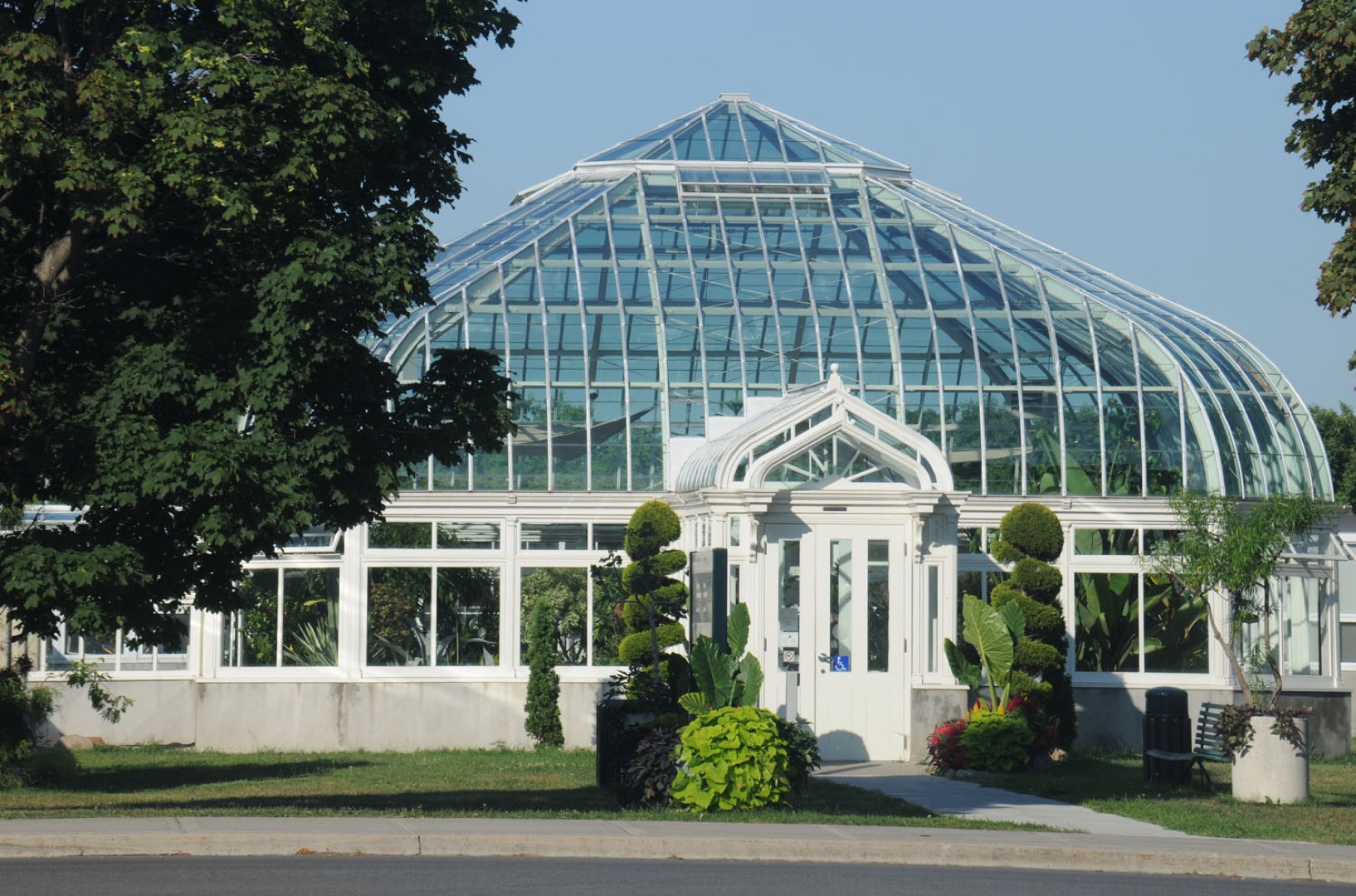 Ici, un bâtiment muni d’une voûte de verre comme toit occupe le centre de la photographie. La porte blanche du bâtiment est entourée d’arbustes verts. À travers le verre, on voit des plantes vertes de différentes tailles. Au premier plan, des branches d’arbres feuillus entrecoupent la photographie.
