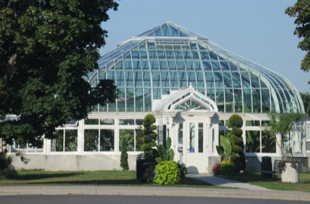 Ici, un bâtiment muni d’une voûte de verre comme toit occupe le centre de la photographie. La porte blanche du bâtiment est entourée d’arbustes verts. À travers le verre, on voit des plantes vertes de différentes tailles. Au premier plan, des branches d’arbres feuillus entrecoupent la photographie.