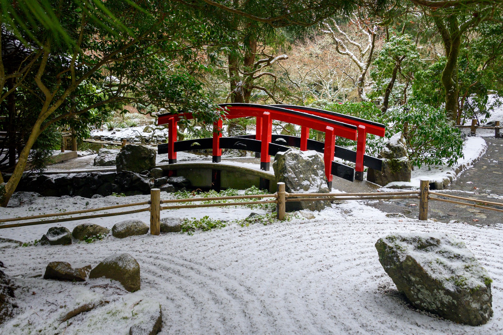Sur cette photographie couleur, le jardin est recouvert d’une fine couche de neige, entre autres, les sculptures de roches et les platebandes du premier plan. À l’arrière-plan, le pont qui prolonge le sentier attire le regard. Au fond, quelques arbres sont dénudés, mais certains ont encore des feuilles.