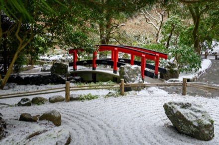 Sur cette photographie couleur, le jardin est recouvert d’une fine couche de neige, entre autres, les sculptures de roches et les platebandes du premier plan. À l’arrière-plan, le pont qui prolonge le sentier attire le regard. Au fond, quelques arbres sont dénudés, mais certains ont encore des feuilles.